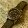 Montre militaire vintage à cadran noir avec bracelet en cuir, posée sur une carte ancienne à côté d’une boussole.